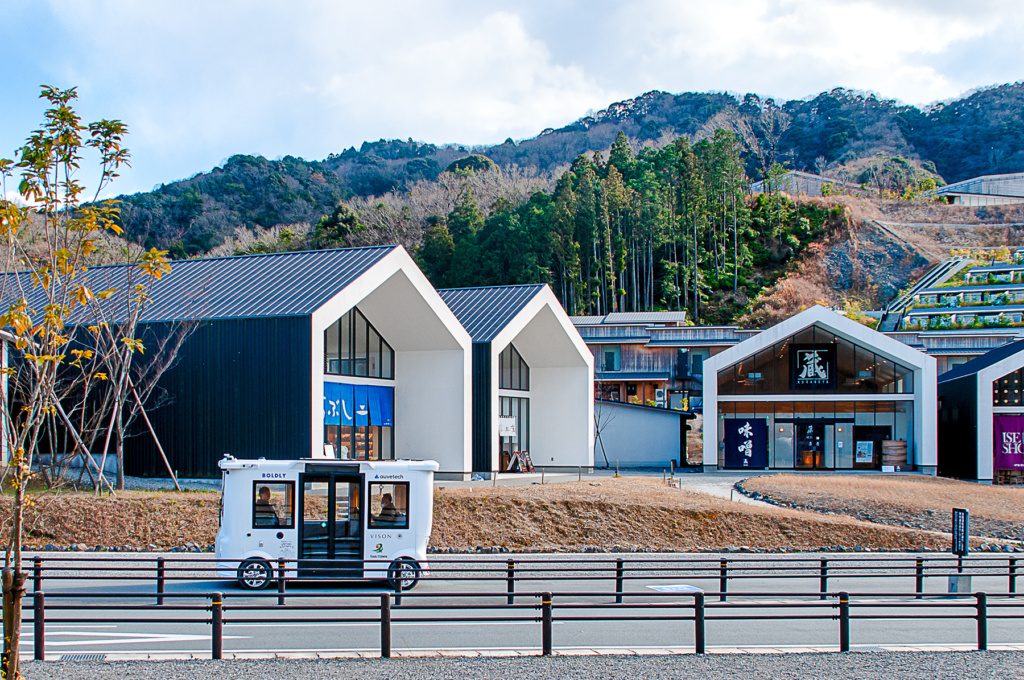 Auve Tech autonomous shuttle operated by Boldly driving in front of modern buildings with forested hills in the background in Japan