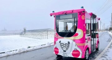 Pink autonomous bus driving on a wet snowy road in Japan with greenhouses in the background