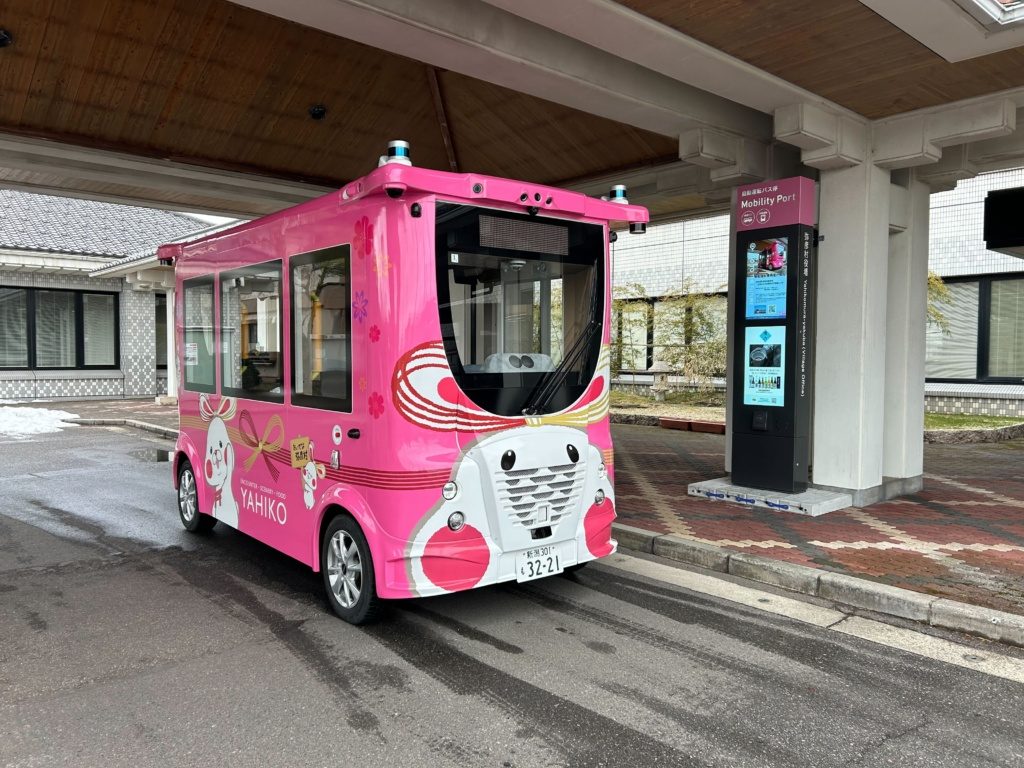 Pink autonomous shuttle in Japan parked at a mobility port with a digital display in the background