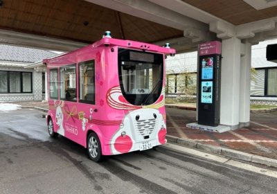 Pink autonomous shuttle in Japan parked at a mobility port with a digital display in the background