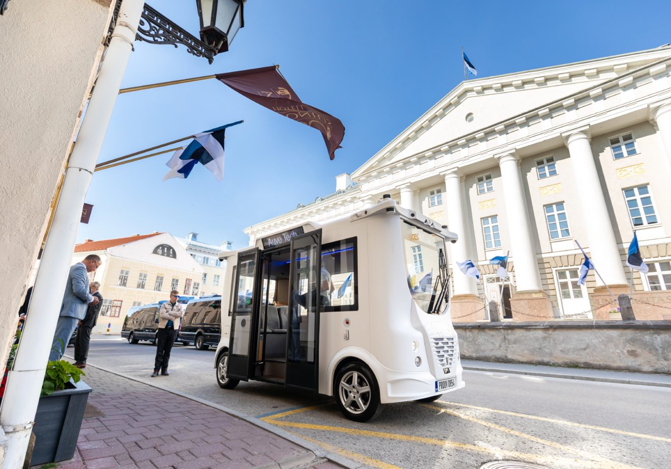 Autonomous shuttle parked on a street in front of a historic building with flags in the background
