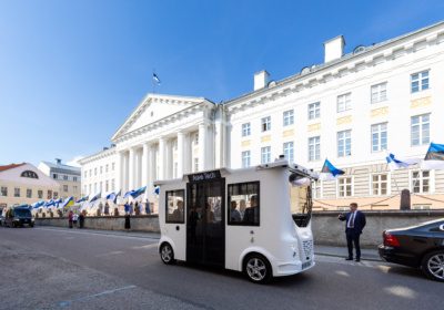 Auve Tech autonom shuttle driving past Tartu University building in Estonia, with Estonian flags displayed along the street