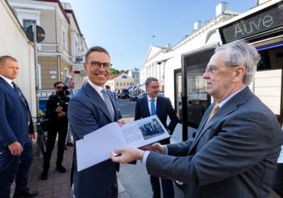 Alexander Stubb receiving a document from Väino Kaldoja next to an autonomous shuttle on a sunny day