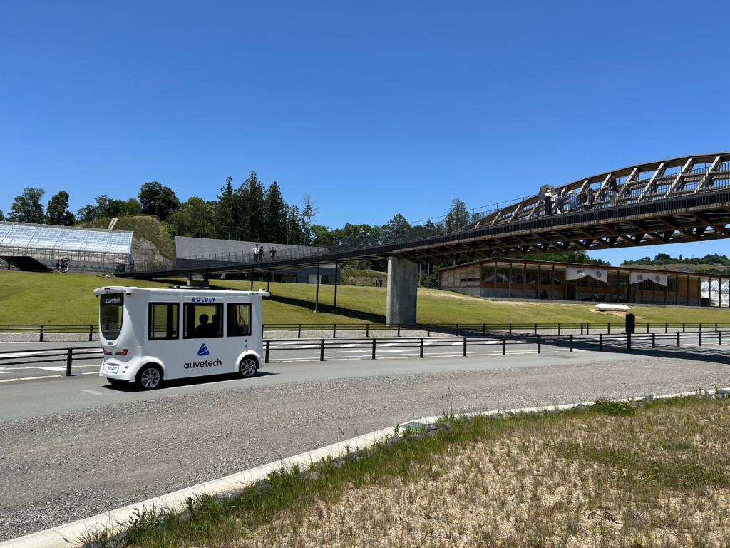 Driverless shuttle by Auve Tech operating at Vision Resort in Japan, near a modern bridge and green landscape.