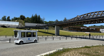 Driverless shuttle by Auve Tech operating at Vision Resort in Japan, near a modern bridge and green landscape.