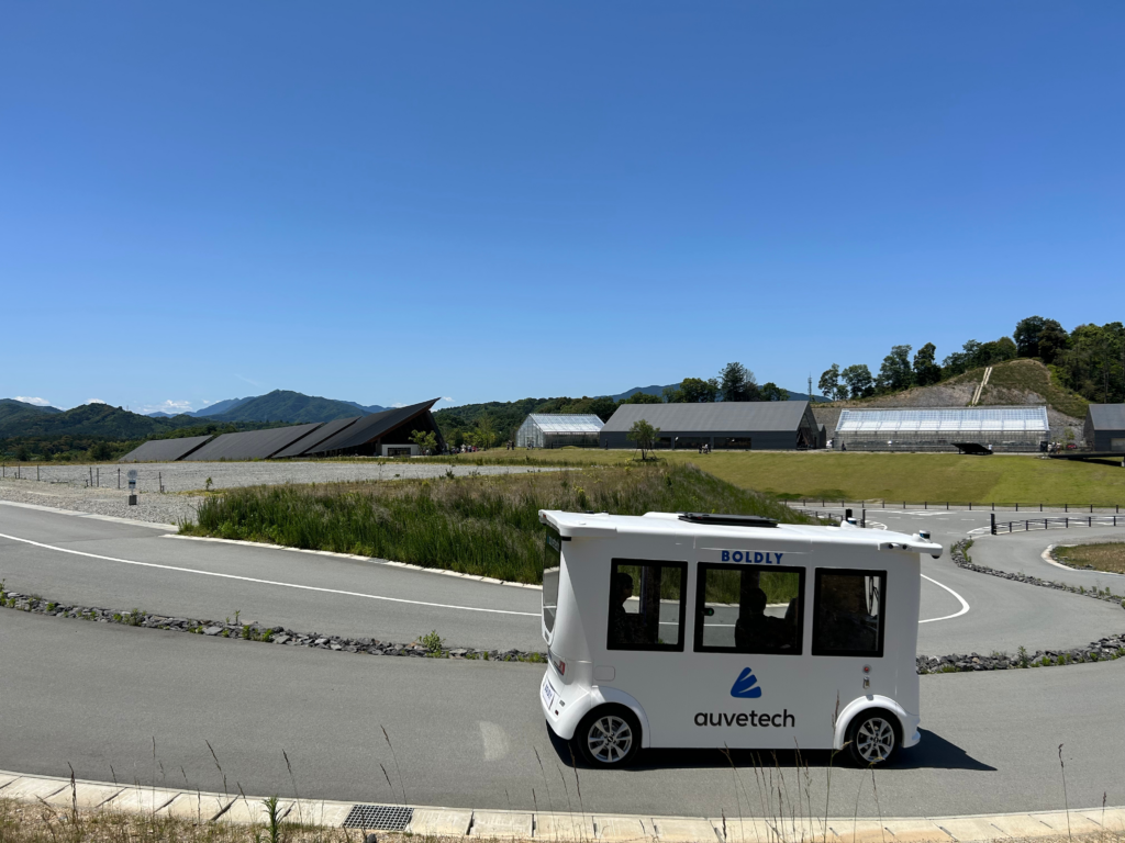 Driverless shuttle by Auve Tech at Vision Resort in Japan, driving on a curved road surrounded by green hills and modern architecture.