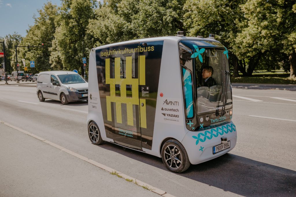 Autonomous shuttle bus in Tartu, Estonia, driving through a sunny urban street with trees in the background, representing modern autonomous transportation.