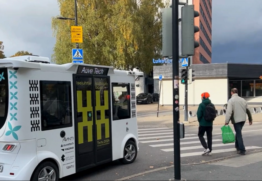 Auve Tech autonomous transportation shuttle MiCa driving near a pedestrian crosswalk on an urban street in Tartu, Estonia, with pedestrians walking and a building in the background.