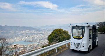 Self-driving bus operating on a scenic hillside road in Takamatsu, Japan, overlooking the city and coastline.